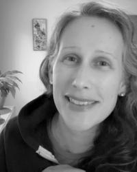 A headshot of me smiling in my home office, in black and white, wearing a hoodie sweatshirt with my plants visible behind me.