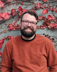 Profile photo of Wilson Beebe. Has beard and glasses and is wearing burnt orange sweater against a backdrop of red leaves.
