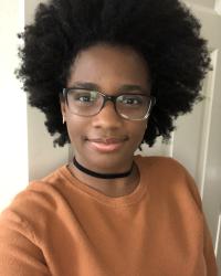 A young Black woman smiling at the camera. She's wearing an orange sweater and black eye glasses.