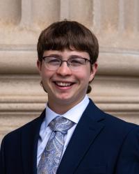 A white person with short hair and glasses wearing a suit and tie smiles at the camera.